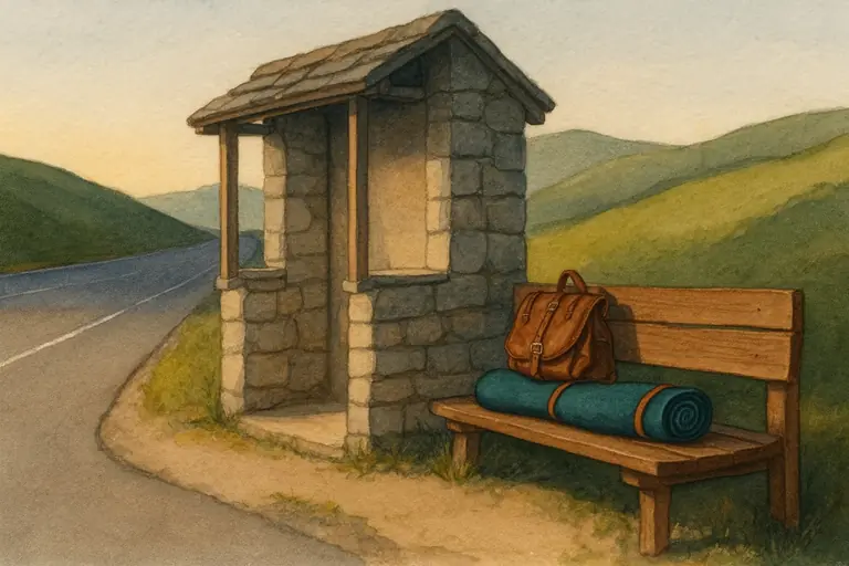 A leather travel bag and a rolled prayer mat on the bench of a stone roadside shelter beside a winding country road.